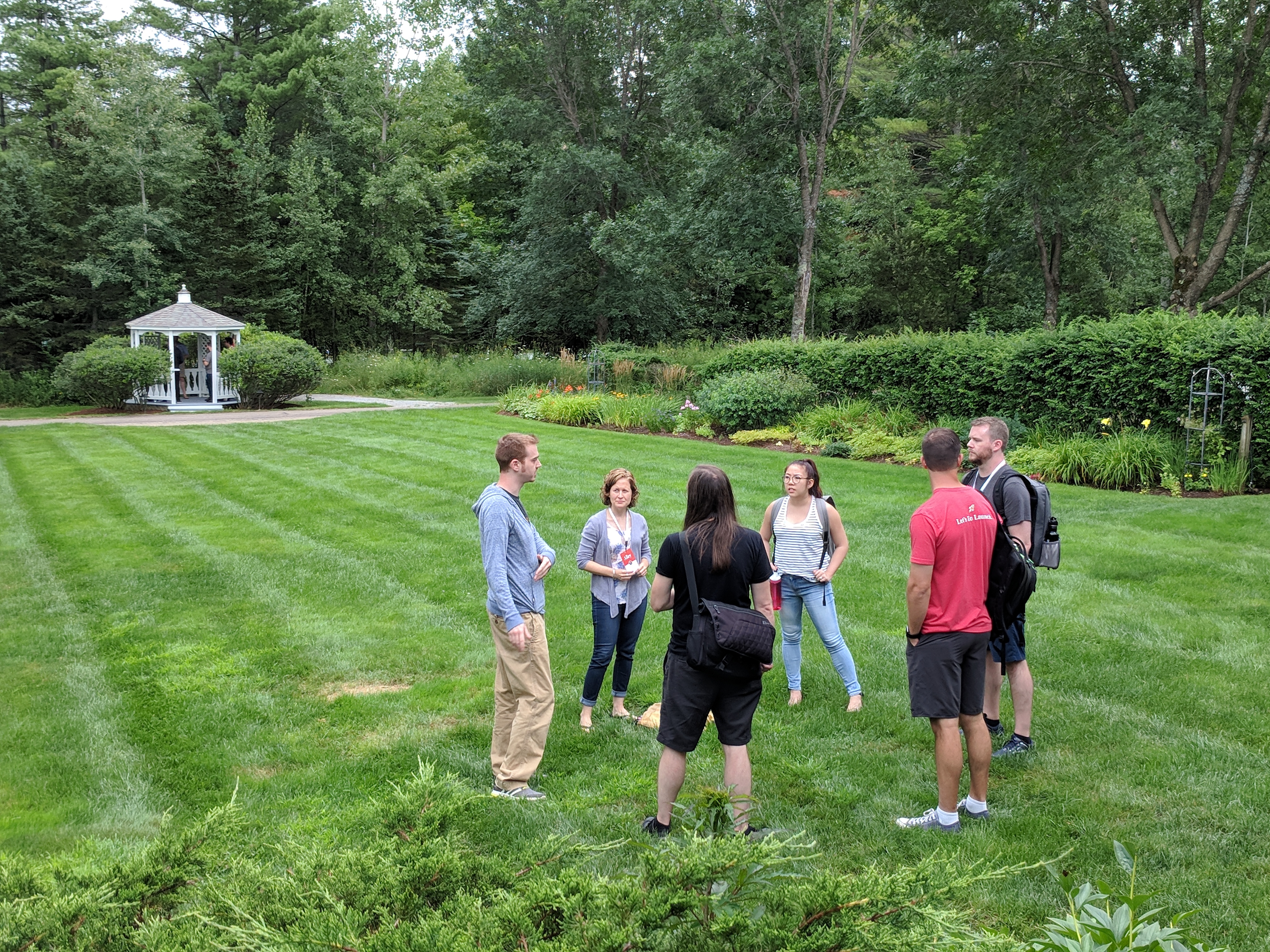 A group of Alley team members standing in a circle at one end of a long green field with a gazebo in the background