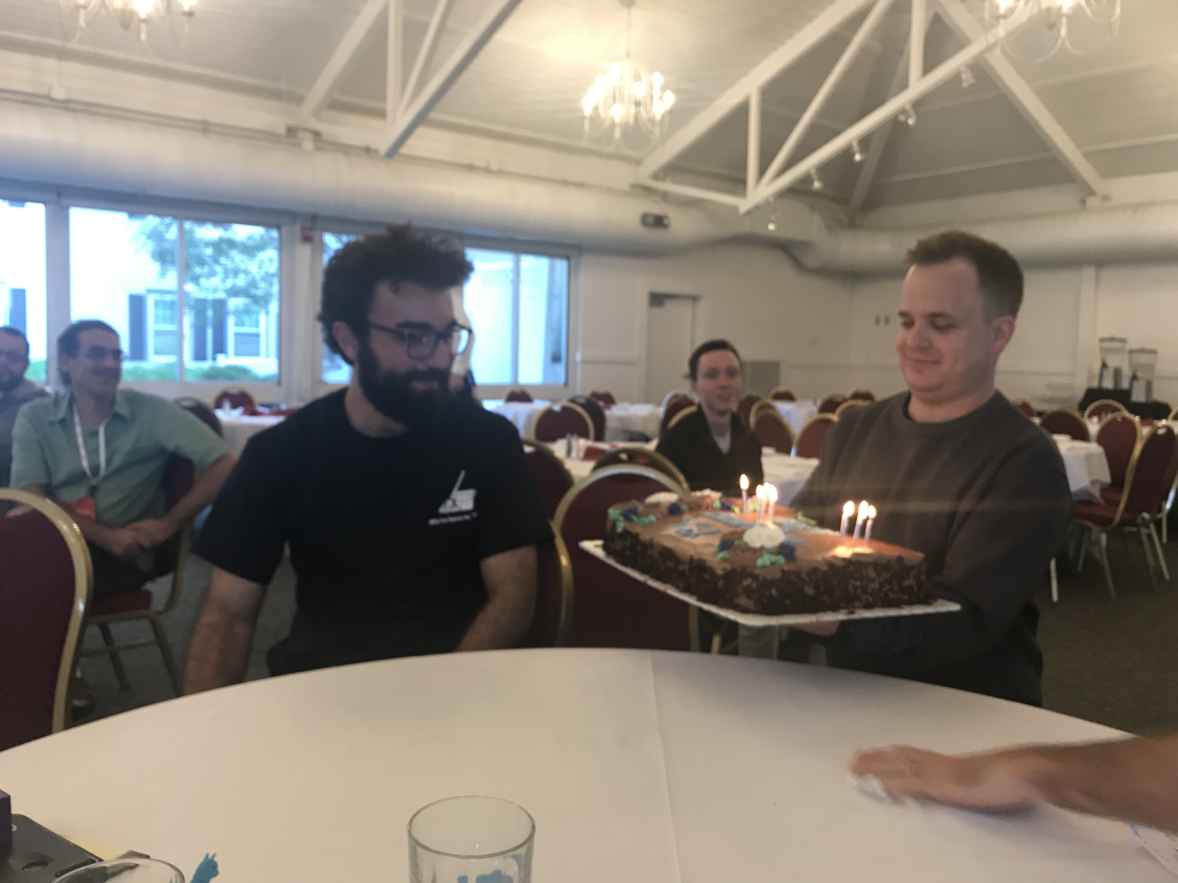 A White man holding a brown cake with lit candles in it and presenting it to a White bearded man with glasses