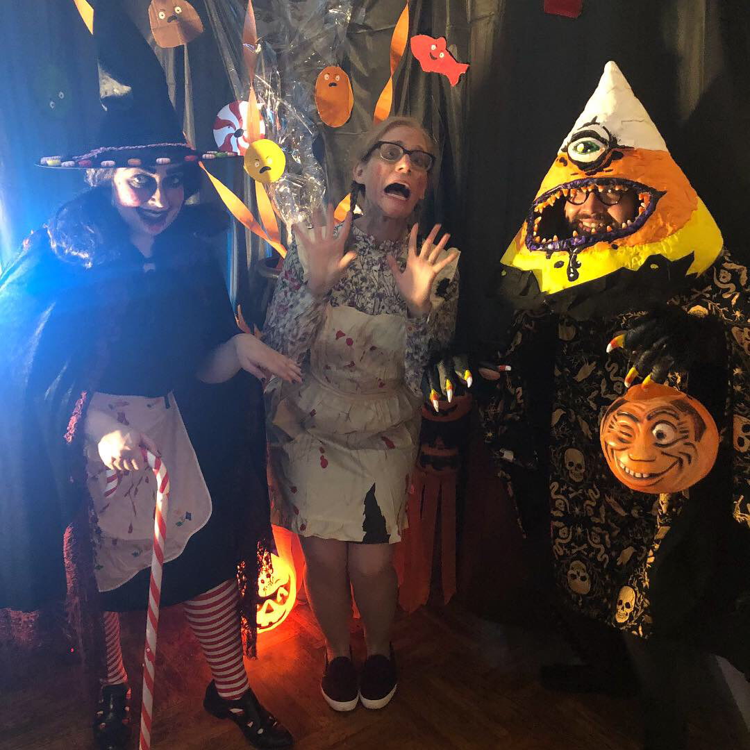 A Halloween party pose with a witch, what appears to be a baker looking scared, and a giant candy-corn monster with teeth and one big eye